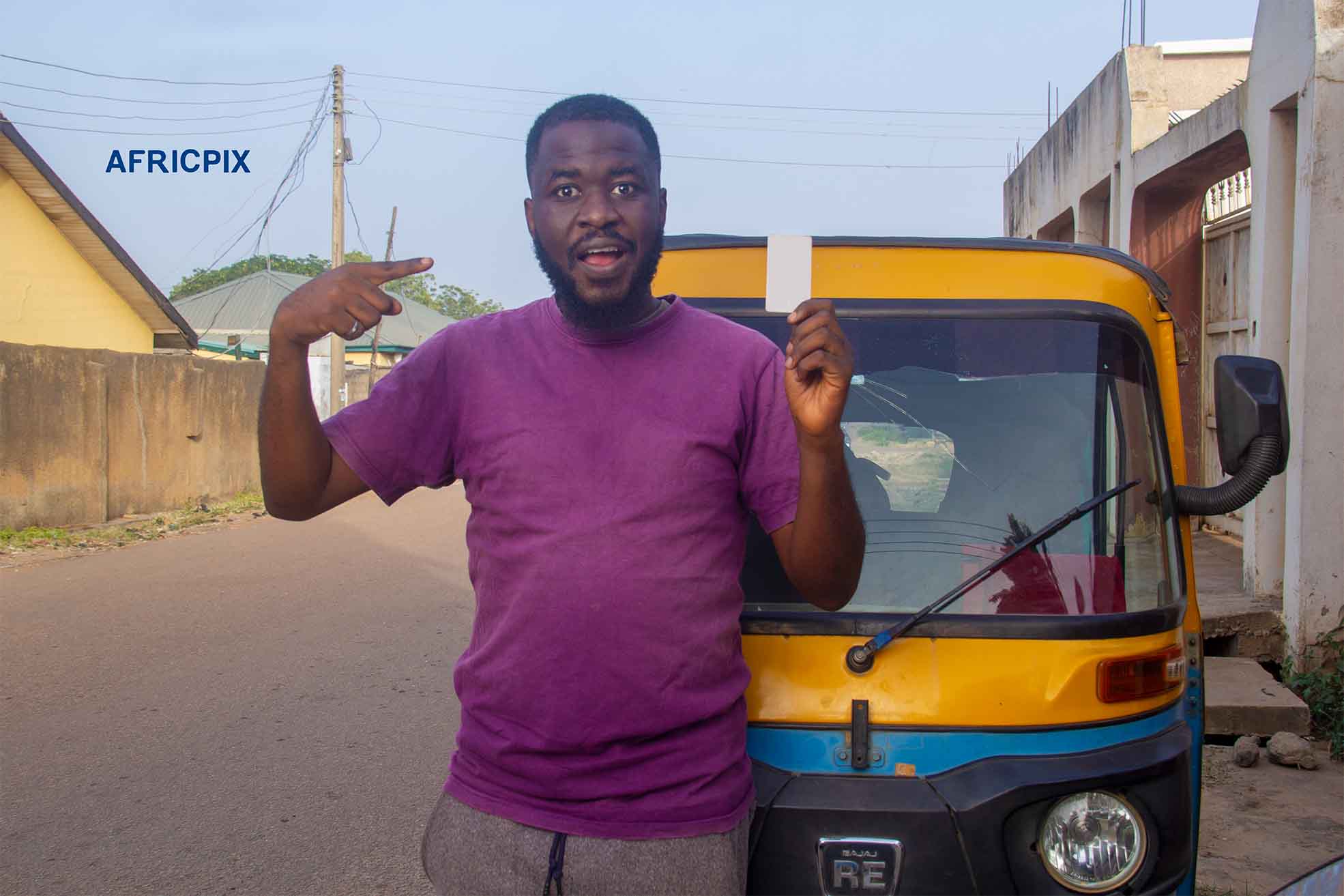 A Surprise, happy African tricycle rider, also known as Keke or Maruwa rider, standing with his tricycle behind him, pointing at debt card, ID card with a cheerful expression.2